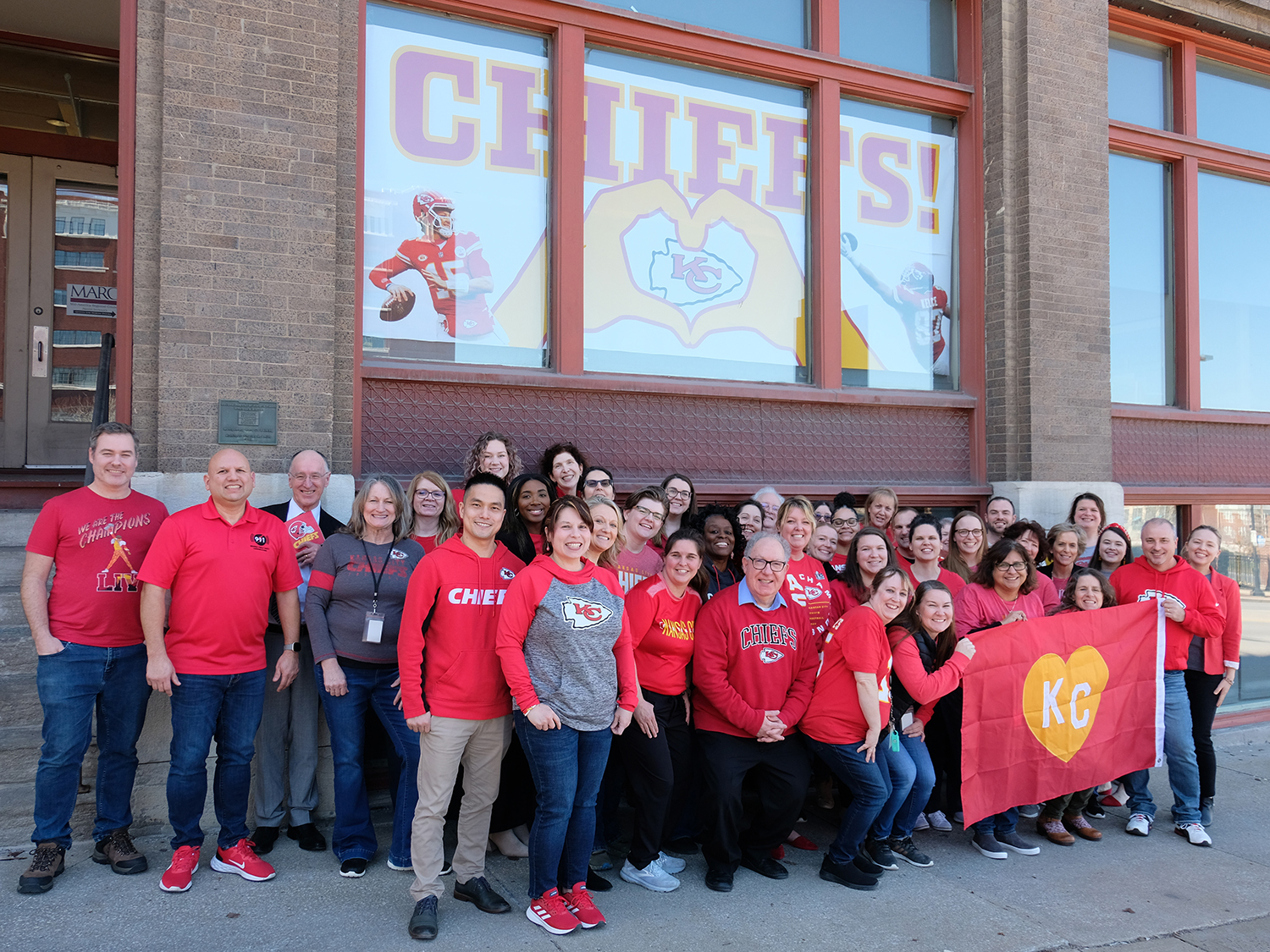 A large group of adults wearing Chiefs attire standing outside holding a red KC flag. 