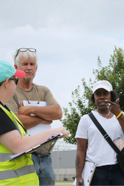 Participants ask questions to training coordinator carrying clipboard in a forested field