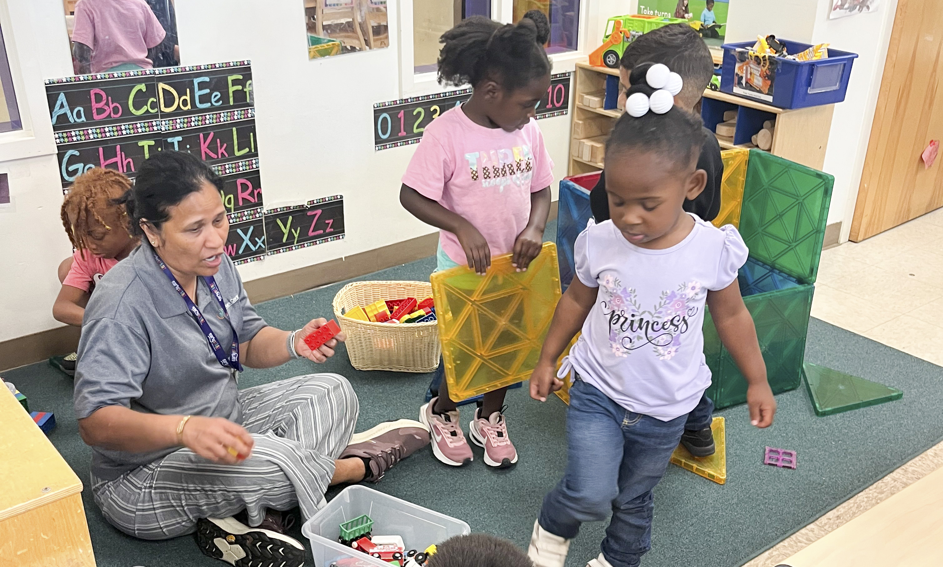 A teacher in a Head Start classroom sits on the floor as students put away blocks