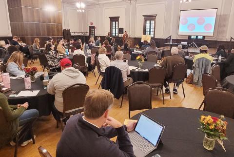 Room of people sitting at round tables looking at a presentation screent