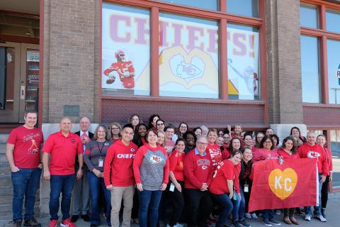 A large group of MARC staff members in red attire standing in front of a Chiefs banner displayed in the window of the MARC offices