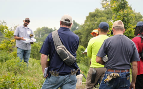 Instructor discusses items on a clipboard to trainees dressed in high-vis clothing in an overgrown field