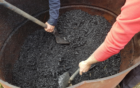 workers mix up black organic material in a bin with shovels