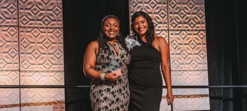 Two women in formal dresses stand on stage and hold awards.