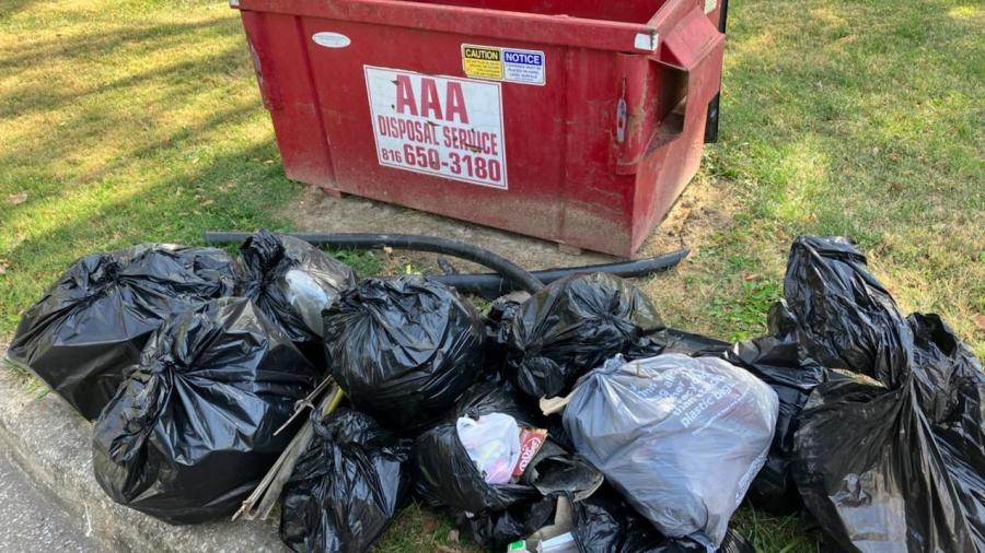 pile of full trash bags outside of a full dumpster along a public park trail