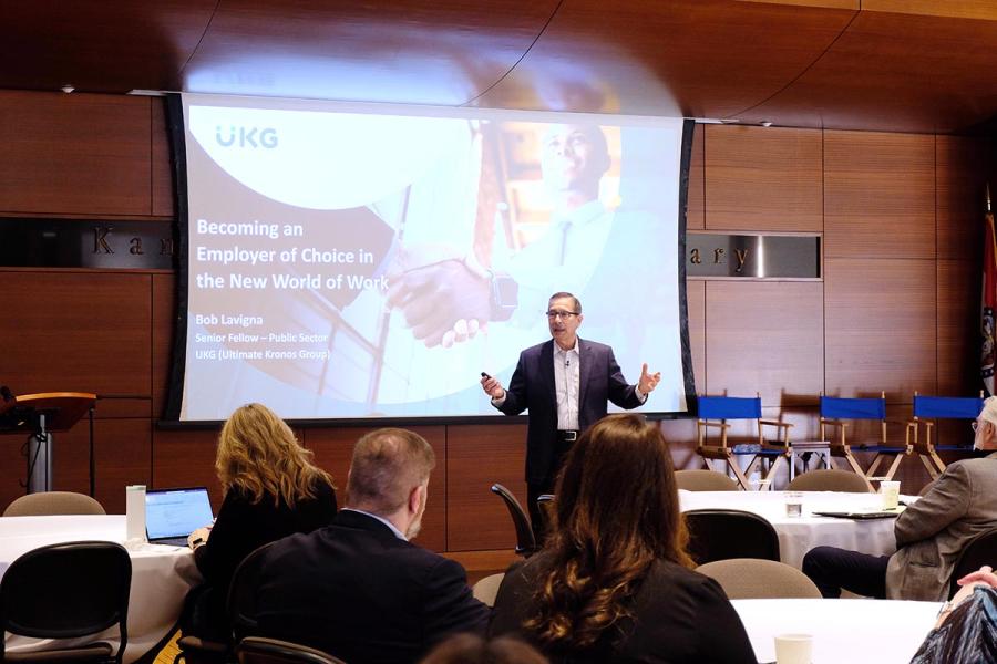 A white man in a suit stands in front of a screen presenting to a room