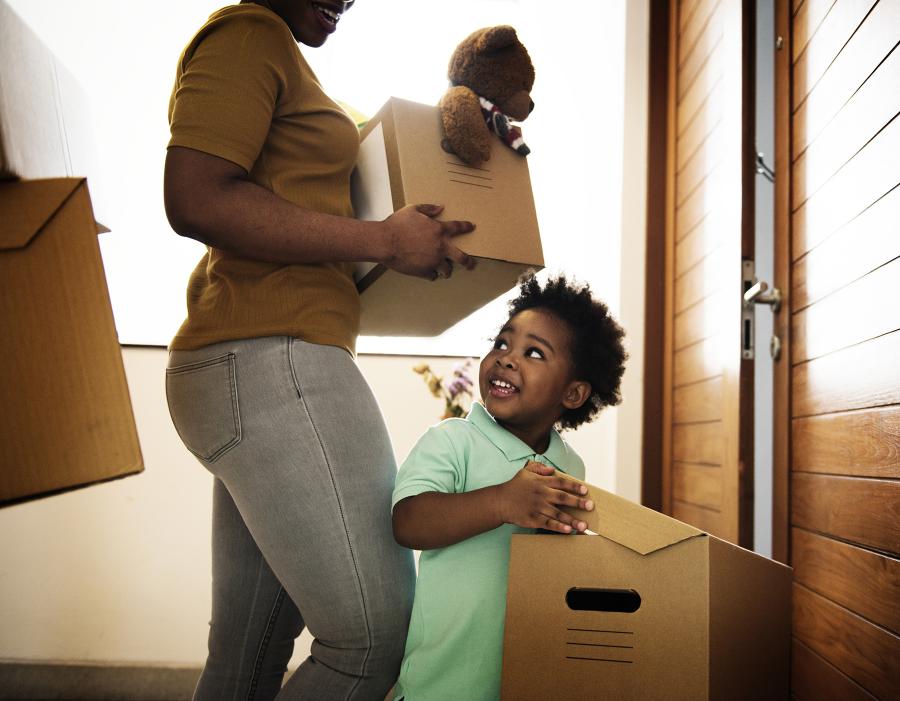 A boy carries a moving box toward a house with his mother close behind, also holding a moving box