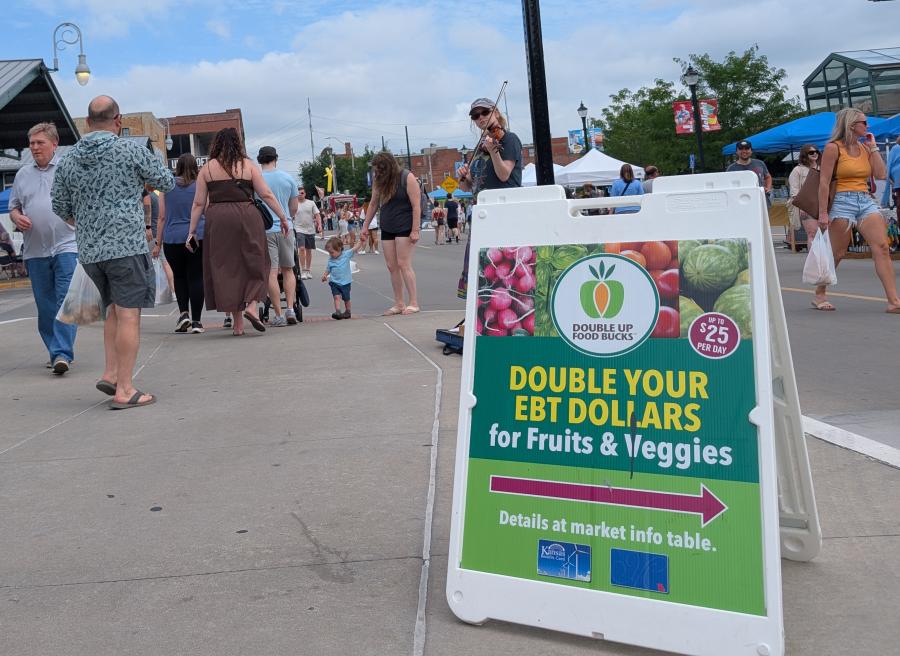 A sign for the Double Up Food Bucks program at the River Market with lots of people around.