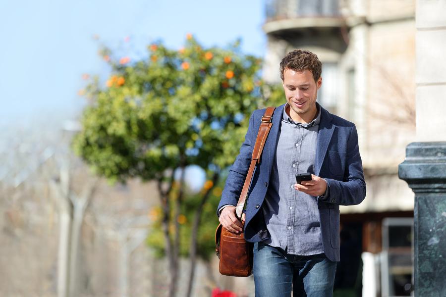 A man walking outside while holding a laptop bag and looking at a cell phone